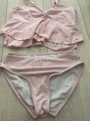 Ljusrosa bikini med volang och broderi - Supersöt ljusrosa bikini med volangdetaljer på toppen och broderade hålmönster över hela setet. Toppen har smala axelband och en liten rosett framtill. Bikinin är fodrad och har en feminin, romantisk stil som passar perfekt till sommaren.