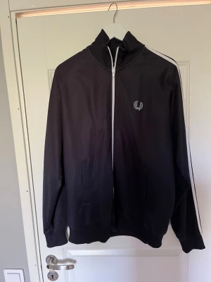 Fred Perry zip hoodie - Snygg svart track jacket med vita ränder längs ärmarna och vit dragkedja. (Fredd Perry). Jackan har hög krage, två fickor framtill och en broderad logga på bröstet. Perfekt för en sportig och avslappnad stil. Koftan är sparsamt använd och i mycket bra skick. Passar både XL och L. PRIS KAN DISKUTERAS.