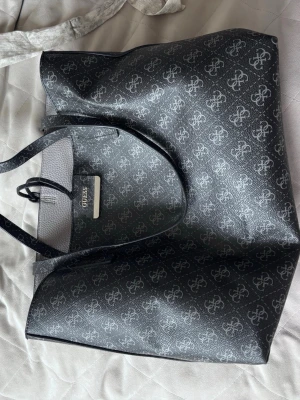 Svart shoppingväska från Guess - Snygg svart shoppingväska från Guess med ikoniskt logomönster över hela väskan. Väskan har dubbla handtag, öppen topp och en praktisk innerficka med dragkedja. Tillverkad i konstläder med silverfärgad logodetalj framtill.