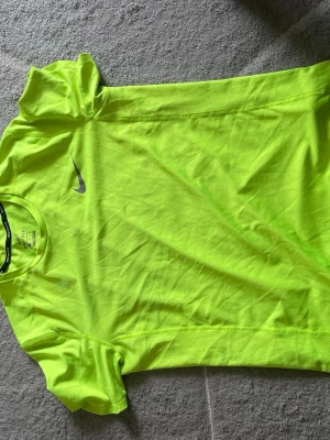 Neongul Nike Dri-FIT löpartröja - Snygg neongul löpartröja från Nike med Dri-FIT-material som andas och transporterar bort svett. Tröjan har korta ärmar, rund hals och diskret Nike-logga på bröstet. Perfekt för löpning eller träning när du vill synas.