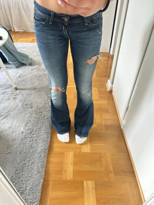 Blå bootcut jeans med slitningar - Säljer ett par blå bootcut jeans med slitningar på båda knäna och lätt tvättade detaljer. Jeansen har klassisk femficksmodell och låg midja. Perfekta för en avslappnad och trendig look. Omgjorda jeans sytt egen bootcut 