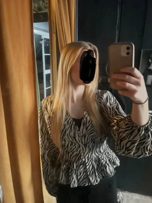 Zebramönstrad blus med volanger - Snygg zebramönstrad blus i svart och vitt med volangdetaljer och v-ringning. Blusen har långa ärmar med resår vid handlederna som ger en puffig effekt. Perfekt för dig som vill sticka ut med ett trendigt djurmönster.