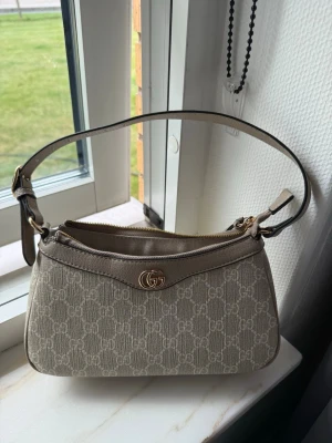 Beige axelremsväska från Gucci - Snygg beige axelremsväska från Gucci med klassiskt GG-mönster i canvas och detaljer i läder. Väskan har guldfärgad logga framtill, justerbar rem och dragkedja upptill. Perfekt för dig som gillar stilrena och trendiga accessoarer. Väskan har varit väll omhändertagen så inga defekter alls. Använd max 4 gånger 