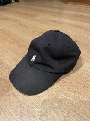 Svart keps från Polo Ralph Lauren - Snygg svart keps från Polo Ralph Lauren med klassisk böjd skärm och justerbart spänne bak. Framtill finns den ikoniska vita polospelaren broderad och baktill står det 'POLO' i vitt. Tillverkad i bomull för en bekväm känsla.