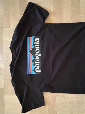 Svart Patagonia t-shirt med tryck - Snygg svart t-shirt från Patagonia med stort färgglatt logotryck på ryggen och mindre logga på bröstet. Klassisk passform och rund halsringning. Tillverkad i mjuk bomull som känns skön mot huden. Perfekt för dig som gillar stilrena och sportiga plagg.