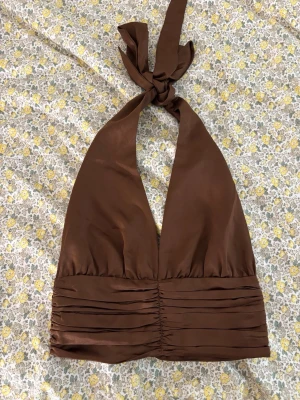 Brun halterneck topp i satin - Snygg brun halterneck topp i satin med bred knytning i nacken och veckade detaljer framtill. Toppen har en croppad passform och knappar i ryggen för en extra cool look. Perfekt för dig som vill sticka ut lite extra i sommar.