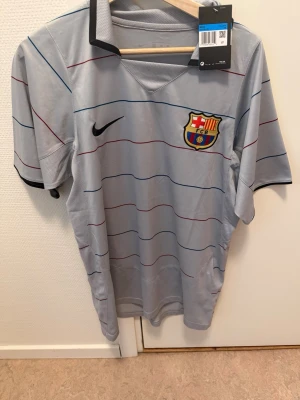 Grå FC Barcelona Ronaldinho tröja - Snygg grå FC Barcelona matchtröja från Nike med tunna ränder i blått och rött. På bröstet finns klubbmärket och Nike-loggan, och på ryggen står det Ronaldinho och nummer 10. Perfekt för fotbollsfans som vill sticka ut på planen eller läktaren.