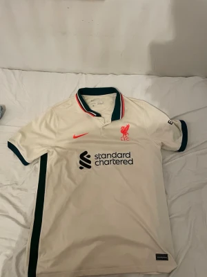 Liverpool andra ställ 2021/22 - Säljer en vit Liverpool matchtröja från Nike med gröna och röda detaljer på kragen. Tröjan har korta ärmar, knappslå och klubbmärke samt sponsortryck på bröstet. Tillverkad i Dri-FIT material som andas, perfekt för träning eller match. Passar M också.