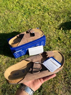 Carafe modell Birkenstock Arizona sandaler - Säljer ett par bruna Birkenstock Arizona sandaler i mocka med två justerbara remmar och metallspännen. Fotbädden är ergonomisk och innersulan är i mjukt material för extra komfort. Perfekta för dig som vill ha både stil och bekvämlighet. Vi snabb affär sänkar jag priset de är riktigt populära så va snabb, säljs billig av en anledning 