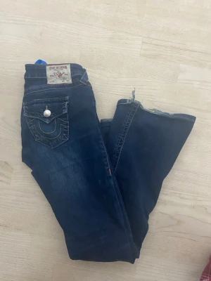 Mörkblå True Religion jeans - Säljer ett par mörkblå jeans från True Religion med klassisk kontrastsöm och snygg fickdetalj med lock och knapp bak. Jeansen har låg midja och raka ben. Perfekta för dig som gillar streetwear och vill ha en cool look.