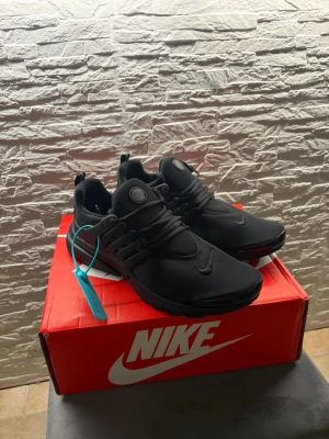Nike Presto svarta sneakers - Snygga helsvarta Nike Presto sneakers med mesh-överdel och plastdetaljer vid snörningen. Skorna har en sportig siluett, rund tå och diskret Nike-logga på sidan och tungan. Perfekta för dig som gillar stilrena och bekväma sneakers.