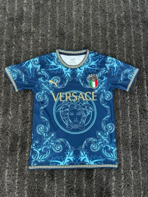 Blå Italien Puma fotbollströja  - Säljer en blå kortärmad fotbollströja med guldiga detaljer och stort Medusa-tryck framtill. T-shirten har ett unikt barockinspirerat mönster i ljusblått och guldfärgade kanter vid hals och ärmslut. Perfekt för dig som vill sticka ut!