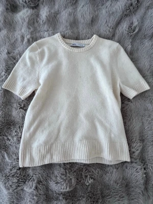 Vit stickad topp från Zara - Säljer en enkel och stilren vit stickad topp från Zara. Toppen har korta ärmar, rund halsringning och ribbade detaljer vid ärmslut och nederkant. Perfekt att matcha med jeans eller kjol för en clean look.