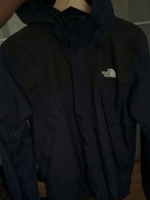 Mörkblå vindjacka från The North Face - Snygg mörkblå vindjacka från The North Face med huva och dragkedja framtill. Jackan har en klassisk logga på bröstet och är tillverkad i ett lätt, vindtätt material som passar perfekt för blåsiga dagar. Pris kan diskuteras.