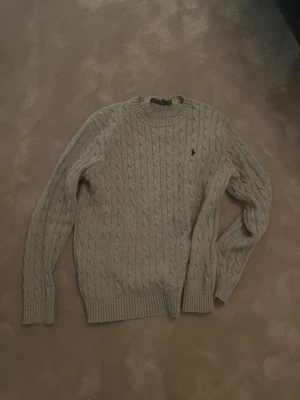 Grå kabelstickad tröja - Kabelstickad tröja i grå med rund halsringning och ribbade muddar vid ärmslut och nederkant. Tröjan har en liten broderad logga på bröstet och är perfekt för lager på lager under kyliga dagar.