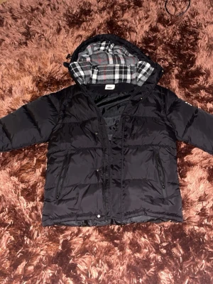 Svart pufferjacka från Burberry - Säljer en svart pufferjacka från Burberry med klassiskt rutigt foder i huvan och tydlig logga på ärmen. Jackan har dragkedja och tryckknappar framtill samt två stora fickor med dragkedja. Perfekt för kalla vinterdagar och riktigt snygg streetstil.