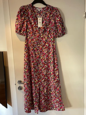 Blommig långklänning från Kappahl - Färgglad långklänning från Kappahl i storlek XS med blommigt mönster i rosa, orange, gul, vit och grön. Klänningen har puffiga korta ärmar och v-ringad hals. Perfekt för sommarens alla tillfällen!
