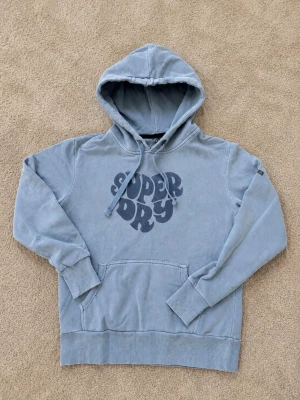 Blå hoodie från Superdry - Passar XS bra. Snygg blå hoodie från Superdry med stor logga i retrostil på bröstet. Hoodien har huva med dragsko och en klassisk magficka. Tillverkad i mjukt bomullsmaterial och har ribbade muddar vid ärmslut och nederkant. Perfekt för en avslappnad look.