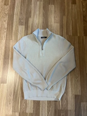 J.Lindberg  halvzip tröja - Snygg beige ribbad tröja med halvzip och hög krage. Perfekt för lager på lager och enkel att matcha med olika outfits. 