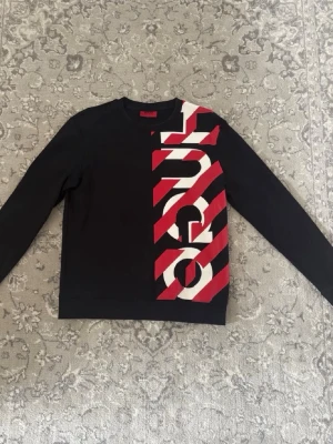 Svart sweatshirt med rödvit text - Säljer en svart sweatshirt med långa ärmar och en stor, vertikal textdetalj i vitt och rött på framsidan. Tröjan har rund halsringning och är gjord i ett mjukt sweatshirtmaterial. Perfekt för dig som gillar streetwear och grafiska tryck.
