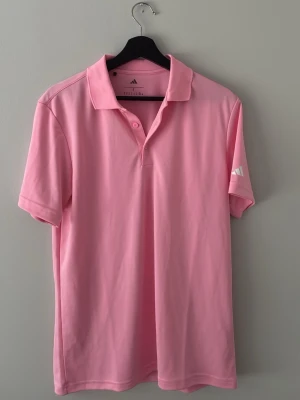 Rosa pikétröja från Adidas helt oanvänd  - Snygg rosa pikétröja från Adidas med klassisk krage och knappar framtill. Tröjan har korta ärmar och Adidas-logga på ärmen. Tillverkad i ett mjukt och lätt syntetmaterial som andas.