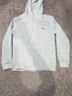 Ljusgrå zip hoodie från Lacoste - Snygg ljusgrå hoodie med dragkedja från Lacoste. Klassisk krokodil-logga på bröstet, huva med dragsko och två fickor framtill. Perfekt för chill dagar eller när du vill ha en clean och enkel stil. Mjuk bomullsblandning för extra komfort.