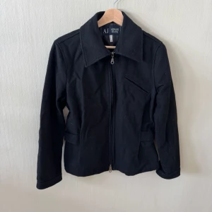 Svart jacka från Armani Jeans - Stilren svart jacka från Armani Jeans med dragkedja framtill, bred krage och fickor med lock. Jackan har justerbara spännen i midjan baktill och en bröstficka med dragkedja. Perfekt för dig som gillar en klassisk look med modern touch.0185
