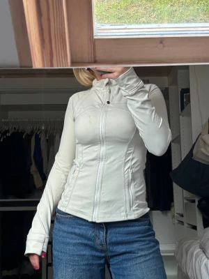 Lululemon Define Jacket - INTRESSEKOLL! Supersnygg vit träningsjacka från Lululemon med figurnära passform, hel dragkedja och två fickor med dragkedja. Jackan har hög krage och långa ärmar med tumhål. Perfekt för dig som vill ha en sportig och clean look. Lite smutsig men tvättas självklart innan jag postar!! Pris kan diskuteras🩷🩷