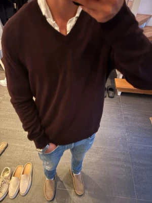 Mörkbrun stickad tröja med V-ringning - Mörkbrun stickad tröja med V-ringning och långa ärmar. Tröjan har en enkel, stilren design och är perfekt att bära över en skjorta för en avslappnad men snygg look. Passar bra till jeans eller chinos. Storlek L passar M