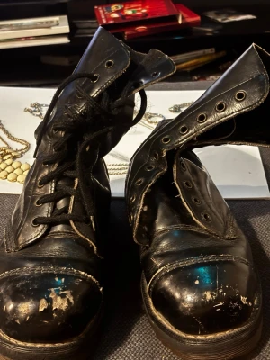 Begagnade Svarta Dr. Martens kängor i läder - Klassiska svarta Dr. Martens kängor i äkta läder med högt skaft och snörning. Ikonisk AirWair-flik baktill och robust sula. Perfekta för dig som gillar en edgy och tidlös stil.