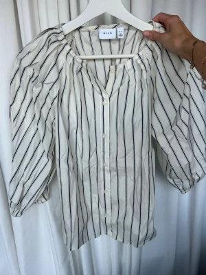 Vit blus med svarta ränder från VILA - Vit blus/skjorta med svarta vertikala ränder från VILA i storlek 34. Puffärm och knappar framtill. Sval och luftig modell, perfekt till vardag eller fest. Använd men i fint skick utan synliga defekter.