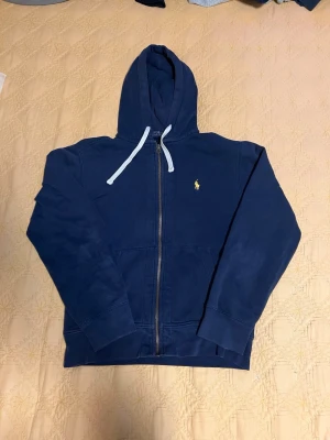 Marinblå hoodie från Polo Ralph Lauren - Snygg marinblå hoodie från Polo Ralph Lauren med dragkedja framtill och vit dragsko i huvan. Klassisk gul broderad logga på bröstet. Tillverkad i mjukt bomullsmaterial och har ribbade muddar vid ärmslut och nederkant. Perfekt för en avslappnad och stilren look.