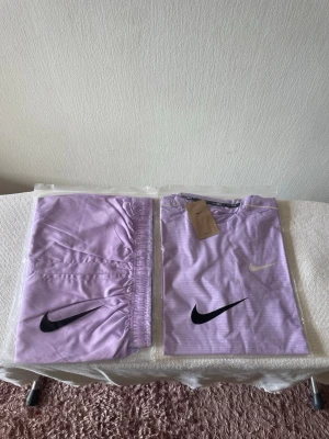 Nike Miller - This image shows a matching Nike sportswear set, consisting of a lavender-colored T-shirt and shorts. Both items are neatly folded and packaged in clear plastic bags. The T-shirt features two Nike swoosh logos (one in black and one in white/grey) on the front, and a Nike tag is attached to the collar. The shorts also display a large black Nike swoosh logo. The set is placed on a white textured surface, likely for display or sale.