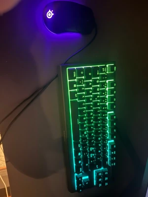 RGB-tangentbord och gamingmus - Säljer ett kompakt mekaniskt tangentbord med RGB-belysning som kan ändra färg mellan grönt, blått och lila. Tangentbordet har svart plastchassi och tydliga vita tecken. Medföljer gör en svart gamingmus med lila LED-belysning och logotyp. Perfekt för gaming eller skrivbordet.