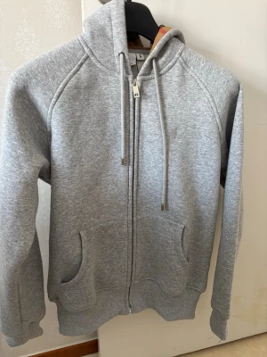 Grå zip hoodie från Burberry - Snygg grå zip hoodie från Burberry med huva och dragsko. Tillverkad i mjukt sweatshirtmaterial och har två fickor framtill samt ribbade muddar vid ärmar och nederkant. Perfekt för en avslappnad och stilren look.