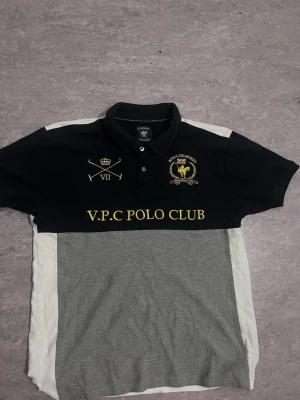 Piké V.P.C Polo Club svart/grå - Snygg pikétröja från V.P.C Polo Club med svart överdel, grå nederdel och vita sidor. Broderade detaljer i gult framtill med text och emblem. Klassisk krage och knappar vid halsen. Perfekt för en sportig och stilren look.
