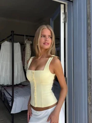 Gul korsett-topp med knappar - Trendig ljusgul korsett-topp med breda axelband och knäppning framtill. Toppen har en figurnära passform och markerad midja, perfekt för att skapa en snygg siluett. Tillverkad i ett mjukt och stretchigt material som känns bekvämt mot huden. Nypris 437 DKK, vilket motsvarar ca 640 KR. Från venderbys, storlek S/M