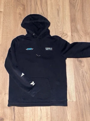 Svart hoodie från Supply & Demand (L) - Svart hoodie med huva och dragsnören från Supply & Demand. Framsidan har liten text och logga, medan baksidan har en stor grafisk ängel med texten 'SUPPLY Demand' i vitt och blått. Fickor framtill och tryck på ena ärmen. Perfekt för en streetwear-look.