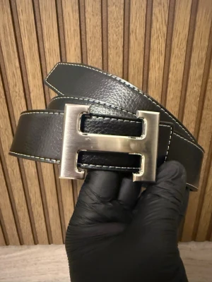 Svart bälte  - Snyggt svart bälte. Bältet har vita kontrastsömmar längs kanterna och en stilren, klassisk look. Perfekt accessoar för att lyfta din outfit.