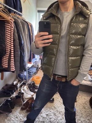 Olivgrön glansig dunväst med huva - Snygg olivgrön dunväst med glansig finish och huva. Västen har dragkedja framtill och två fickor med dragkedja. Perfekt för lager-på-lager och ger en cool streetwear-look.