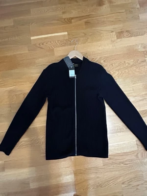 Svart ribbad ziptröja från Gabba - Snygg svart ribbad tröja från Gabba med dragkedja framtill. Tröjan har lång ärm och en stilren, enkel design som passar till det mesta. Tillverkad i ett mjukt stickat material som ger en skön känsla.