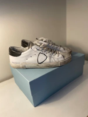 Phillipe modell skor  - Säljer ett par vita sneakers med grå detaljer och svart häl. Skorna har en klassisk låg siluett, vita snören och en dekorativ sköld på sidan. Tillverkade i läder med slitstark sula. Perfekta för en clean och stilren look.