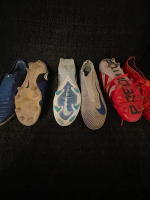 Nike och Adidas fotbollsskor - Fyra par fotbollsskor från Nike och Adidas i olika färger: blå, vit och röd. Skorna har dobbar för spel på gräs och är tillverkade i syntetmaterial. Ett par är Adidas Predator med tydlig logga, övriga är Nike med klassisk swoosh och modern design.