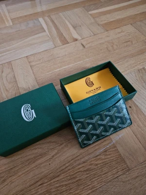 Grön Korthållare - Snyggt grönt kortfodral med klassiskt mönster i vitt och beige. Tillverkad i exklusivt läder med flera fack för kort. Kommer i box och med gul putsduk. Perfekt för dig som vill ha något stilrent och lyxigt i fickan.
