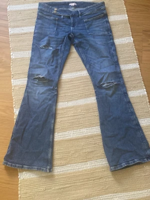 Blå bootcut jeans från Gina Tricot - Säljer ett par blå bootcut jeans från Gina Tricot med slitna detaljer på låren och knäna. Jeansen har låg midja, dubbla knappar och klassiska fickor fram. Perfekta för dig som gillar en retro och avslappnad stil.