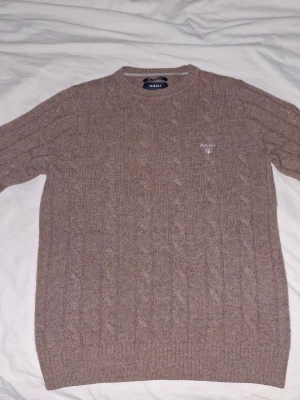 Beige kabelstickad tröja från GANT - Kabelstickad tröja i beige från GANT med rund halsringning och diskret broderad logga på bröstet. Tillverkad i mjuk lammull för extra komfort. Klassisk modell med ribbade muddar vid ärmslut och nederkant.