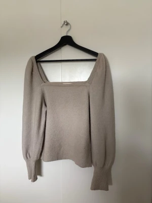 Beige stickad tröja med puffärm - Trendig beige stickad tröja med fyrkantig ringning och puffiga långa ärmar. Tröjan har breda ribbade muddar vid ärmslut och nederkant. Perfekt för dig som gillar en stilren och modern look. Passar även S