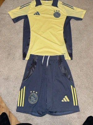 Ajax träningsset från Adidas gul/blå S - Ajax träningsset från Adidas i gul och marinblå. Tröjan har Ajax-logga och Adidas-märke på bröstet, samt tre stjärnor ovanför loggan. Shortsen har dragsko, fickor och matchande detaljer med gula ränder och Ajax-emblem. Materialet är lätt och andas, perfekt för träning.