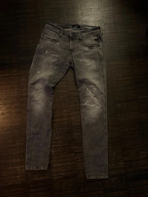 Grå Replay jeans med slitningar - Säljer ett par grå jeans med slitningar och lätt tvättad look. Jeansen har fem fickor och dragkedja i gylfen. Perfekta för dig som gillar en avslappnad men trendig stil.