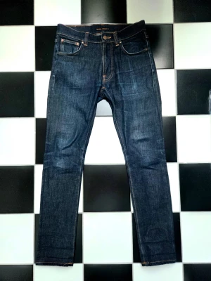 Nudie Jeans Lean Dean - Säljer ett par mörkblå jeans från Nudie Jeans med smal passform och klassiska fem fickor. Jeansen har kontrastsömmar och Nudie Jeans signaturbroderi på bakfickorna. Tillverkade i slitstark denim, perfekta för en clean och stilren look.